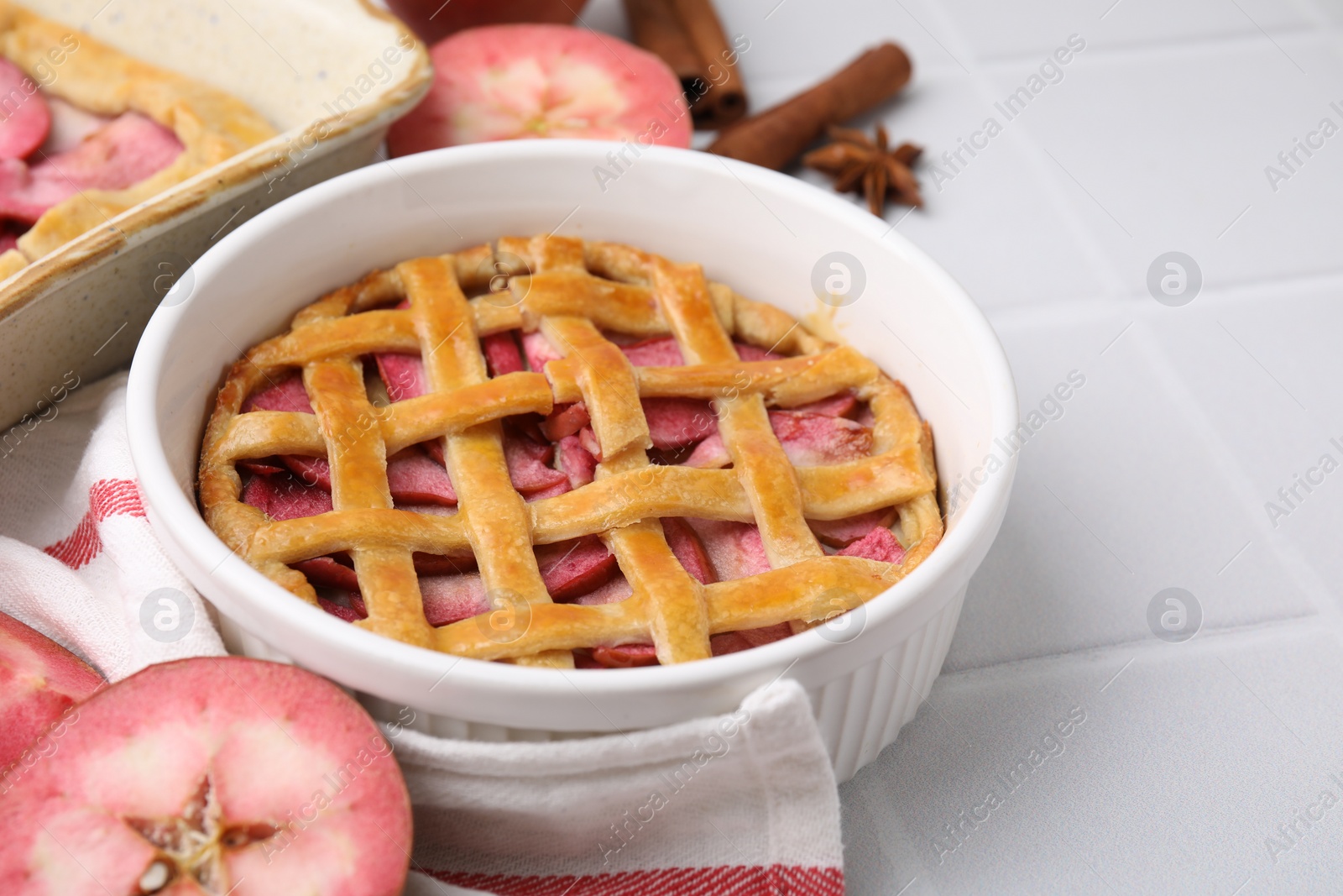 Photo of Baking dish with delicious apple pie and ingredients on white tiled table, closeup