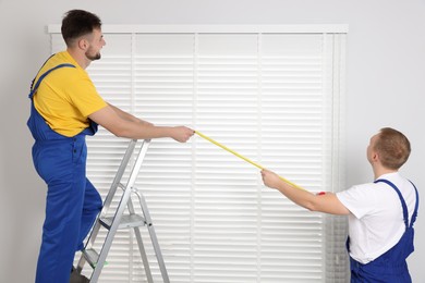 Workers using measuring tape while installing horizontal window blinds indoors