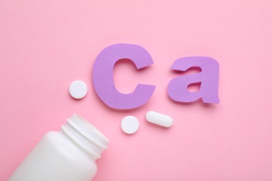 Photo of Pills, open bottle and calcium symbol made of purple letters on pink background, flat lay