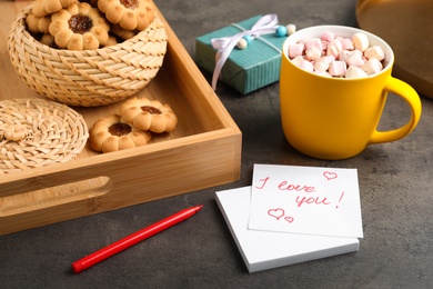 Photo of Note with phrase I Love You near cookies, gift box and cup of hot drink on dark table