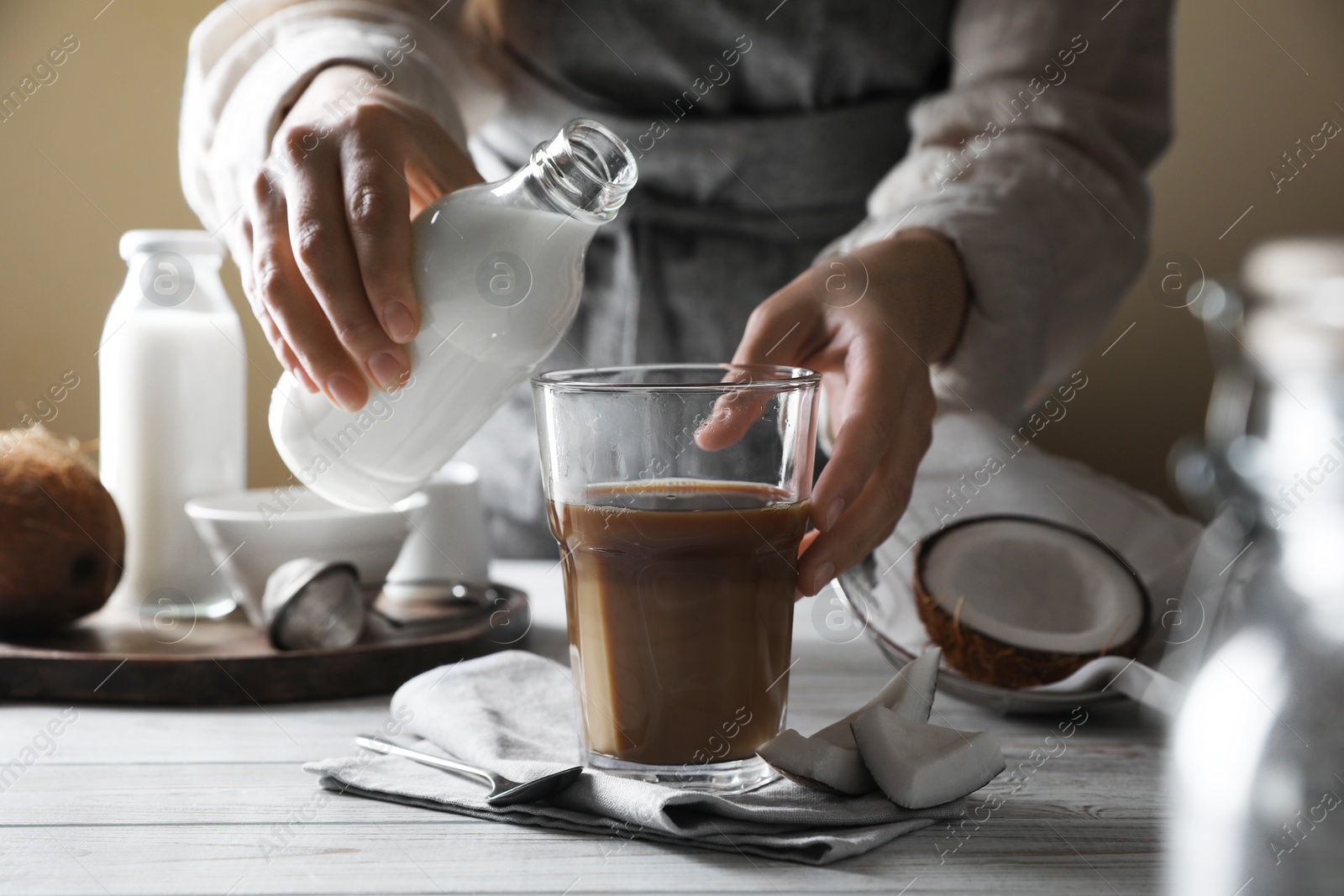 Photo of Woman holding bottle with coconut milk near glass of coffee at white wooden table, closeup