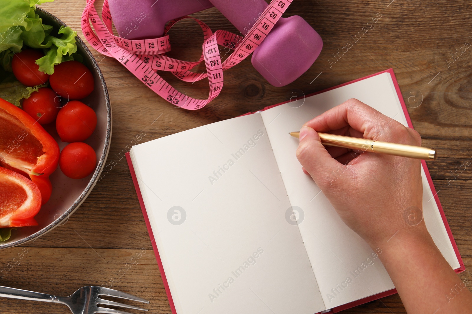 Photo of Woman developing diet plan at wooden table with products, measuring tape and dumbbells, top view