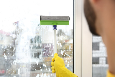 Photo of Professional young janitor in uniform cleaning window indoors, closeup