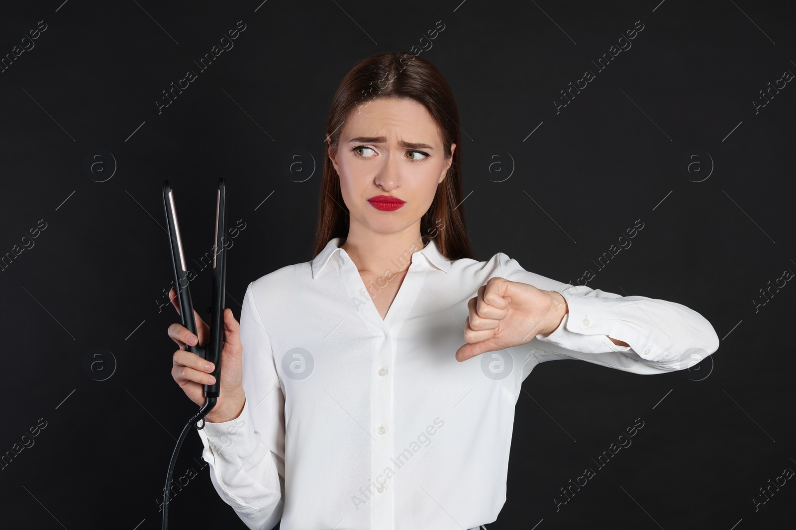 Photo of Upset young woman with flattening iron showing thumb down on black background. Hair damage