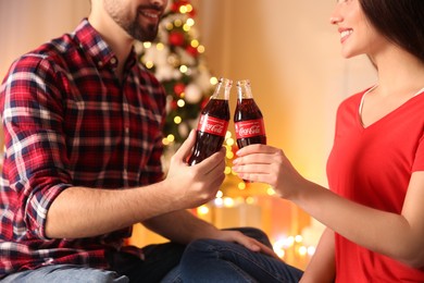 MYKOLAIV, UKRAINE - JANUARY 27, 2021: Young couple holding bottles of Coca-Cola in room decorated for Christmas, closeup