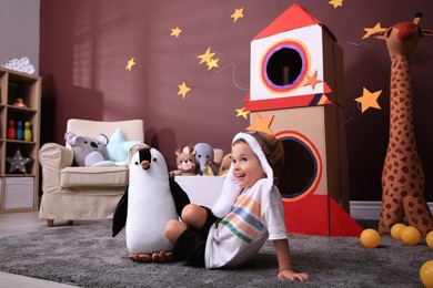 Photo of Cute little boy playing on floor near cardboard rocket  at home. Child's room interior