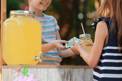 Photo of Little girl selling natural lemonade to kid in park, closeup. Summer refreshing drink