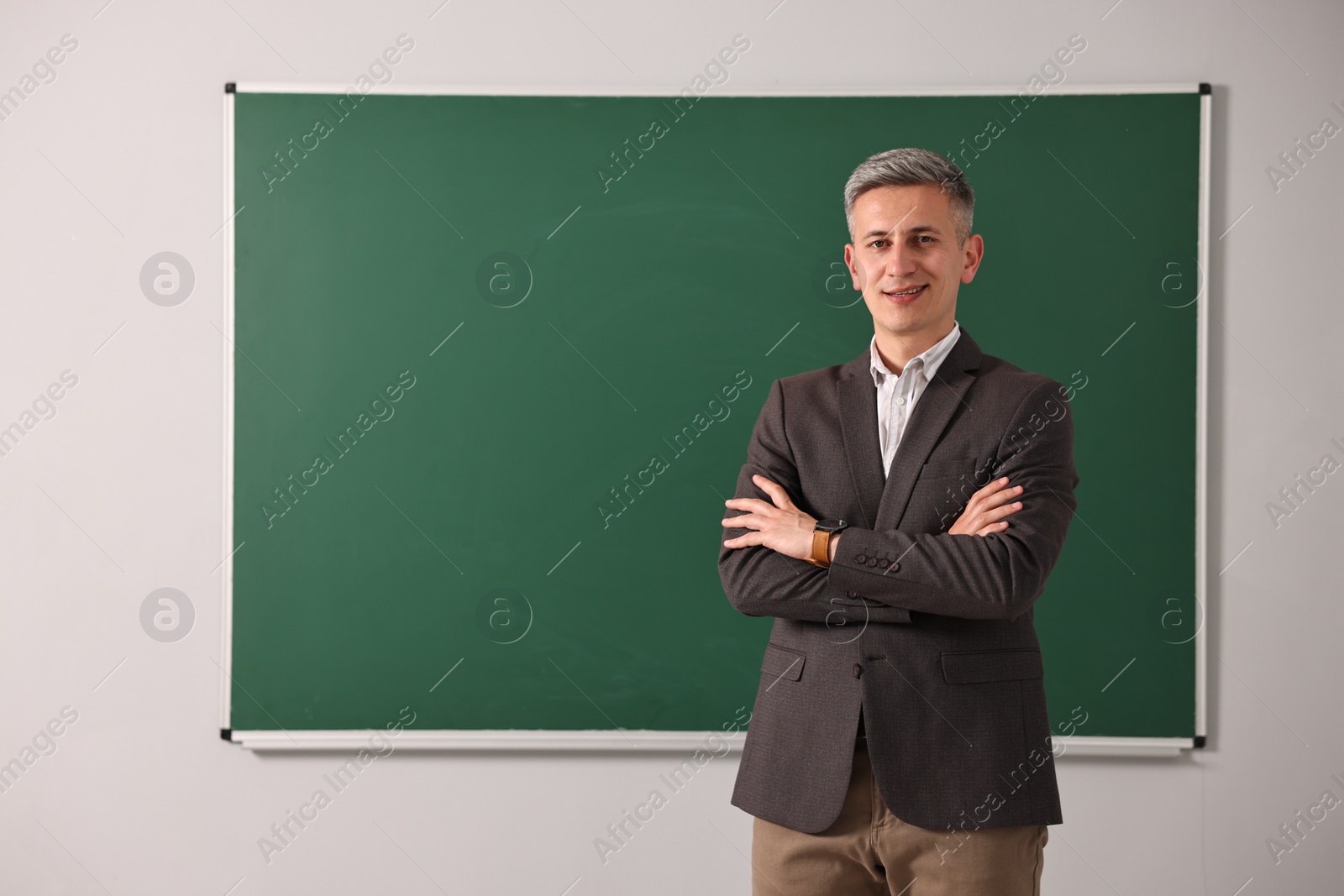 Photo of Happy teacher with crossed arms near chalkboard in classroom, space for text