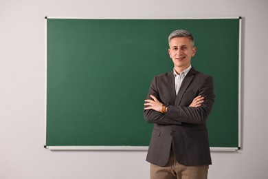 Happy teacher with crossed arms near chalkboard in classroom, space for text