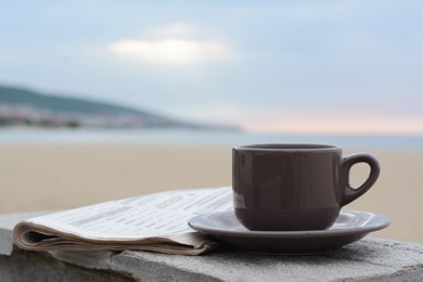 Photo of Ceramic cup of hot drink and newspaper on stone surface near sea in morning