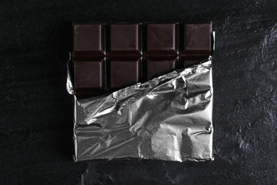 Photo of Tasty dark chocolate bar wrapped in foil on black table, top view