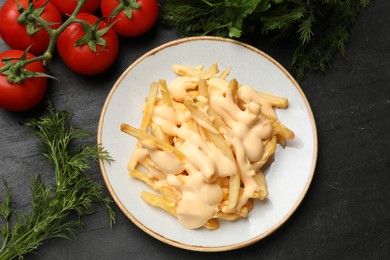 Photo of Delicious French fries with cheese sauce, tomatoes and dill on black table, flat lay