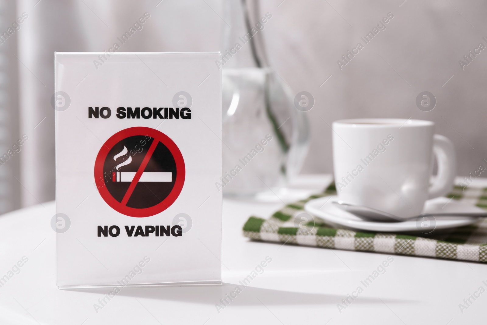 Photo of No Smoking No Vaping sign and cup of coffee on white table indoors