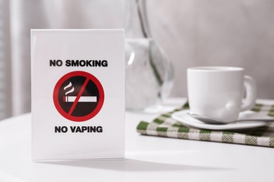 Photo of No Smoking No Vaping sign and cup of coffee on white table indoors