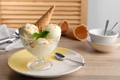 Photo of Delicious scoops of vanilla ice cream with wafer cone and mint in glass dessert bowl on wooden table