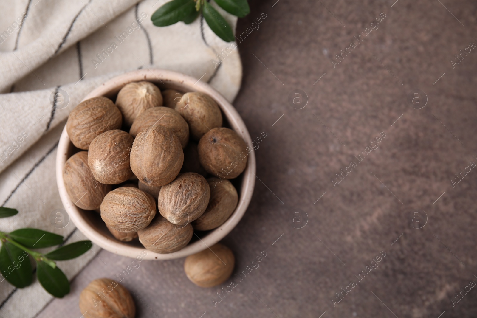 Photo of Whole nutmegs in bowl and green branches on brown table, flat lay. Space for text