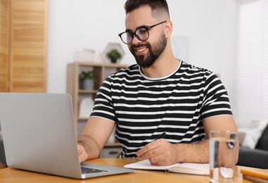 Young man writing in notebook while working on laptop at wooden table indoors