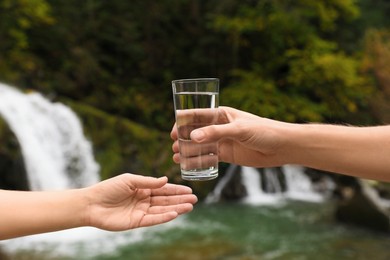 Photo of Man giving woman glass of fresh water near waterfall, closeup