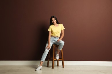 Beautiful young woman sitting on stool near brown wall
