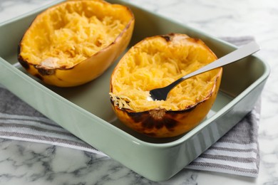 Photo of Halves of cooked spaghetti squash and fork in baking dish on white marble table, closeup