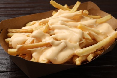 Photo of Tasty potato fries and cheese sauce in paper container on wooden table, closeup