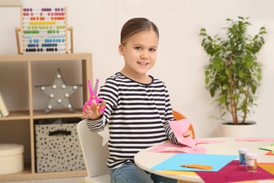 Cute little girl with scissors and colorful paper at desk in room. Home workplace