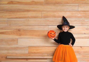 Photo of Cute little girl with pumpkin candy bucket wearing Halloween costume on wooden background. Space for text