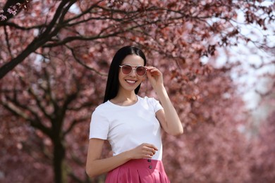 Photo of Pretty young woman with sunglasses near beautiful blossoming trees outdoors. Stylish spring look