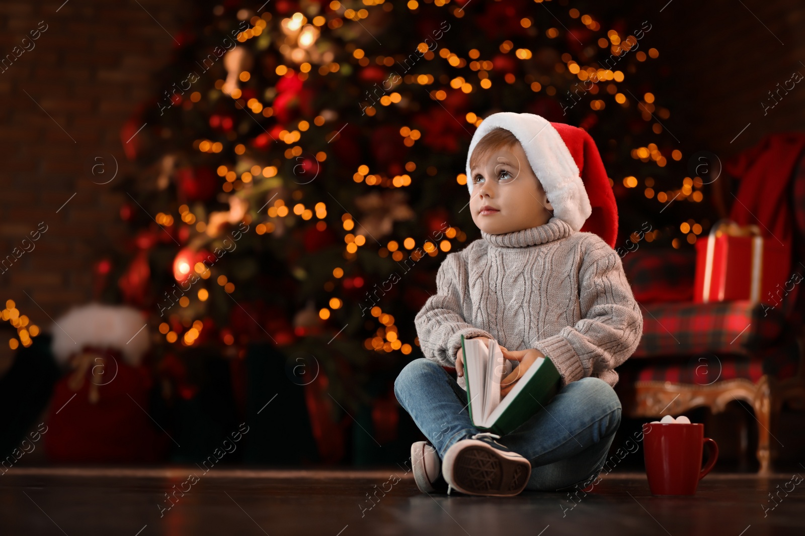 Photo of Little boy in Santa Claus cap with book near Christmas tree at home