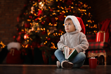 Photo of Little boy in Santa Claus cap with book near Christmas tree at home