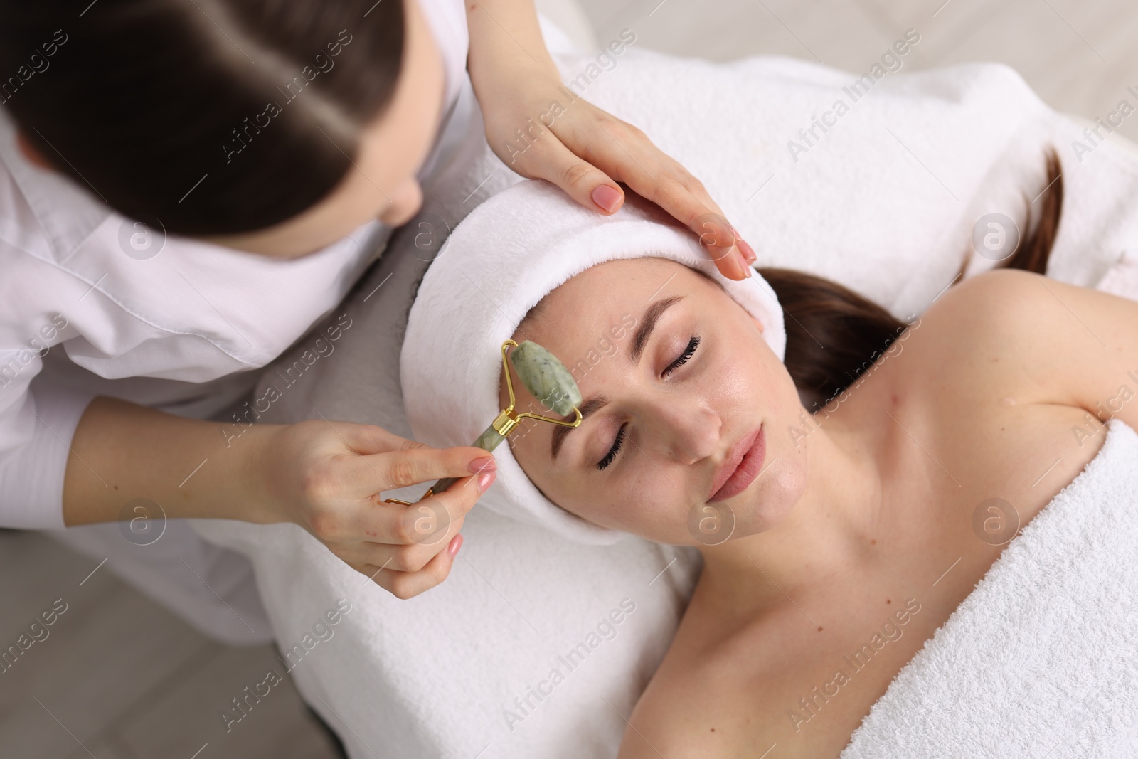 Photo of Cosmetologist making face massage with roller to client in clinic, top view