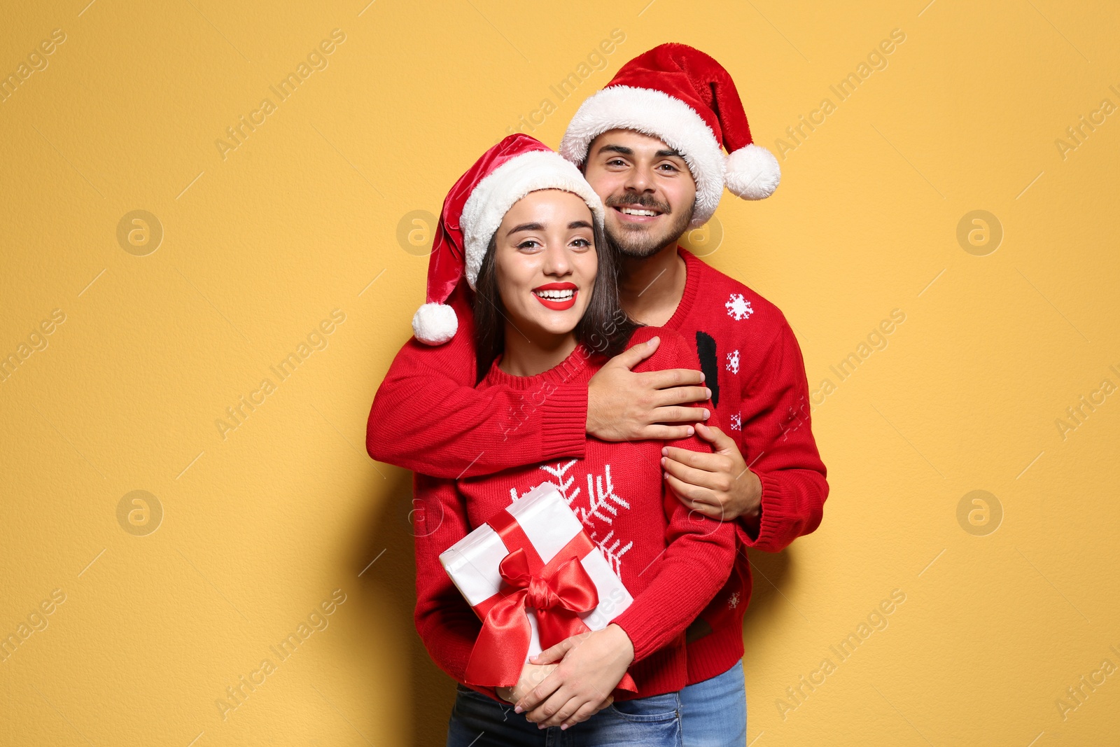 Photo of Young couple in Christmas sweaters and hats with gift on color background
