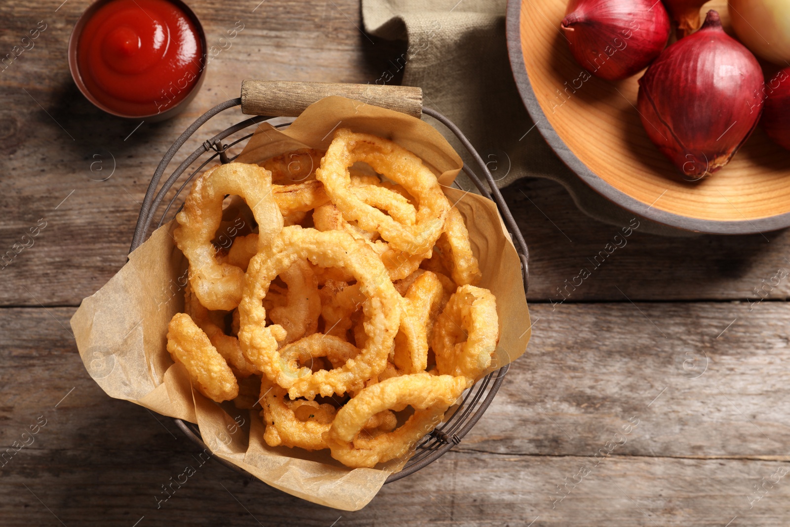 Photo of Homemade crunchy fried onion rings in wire basket on wooden background, top view