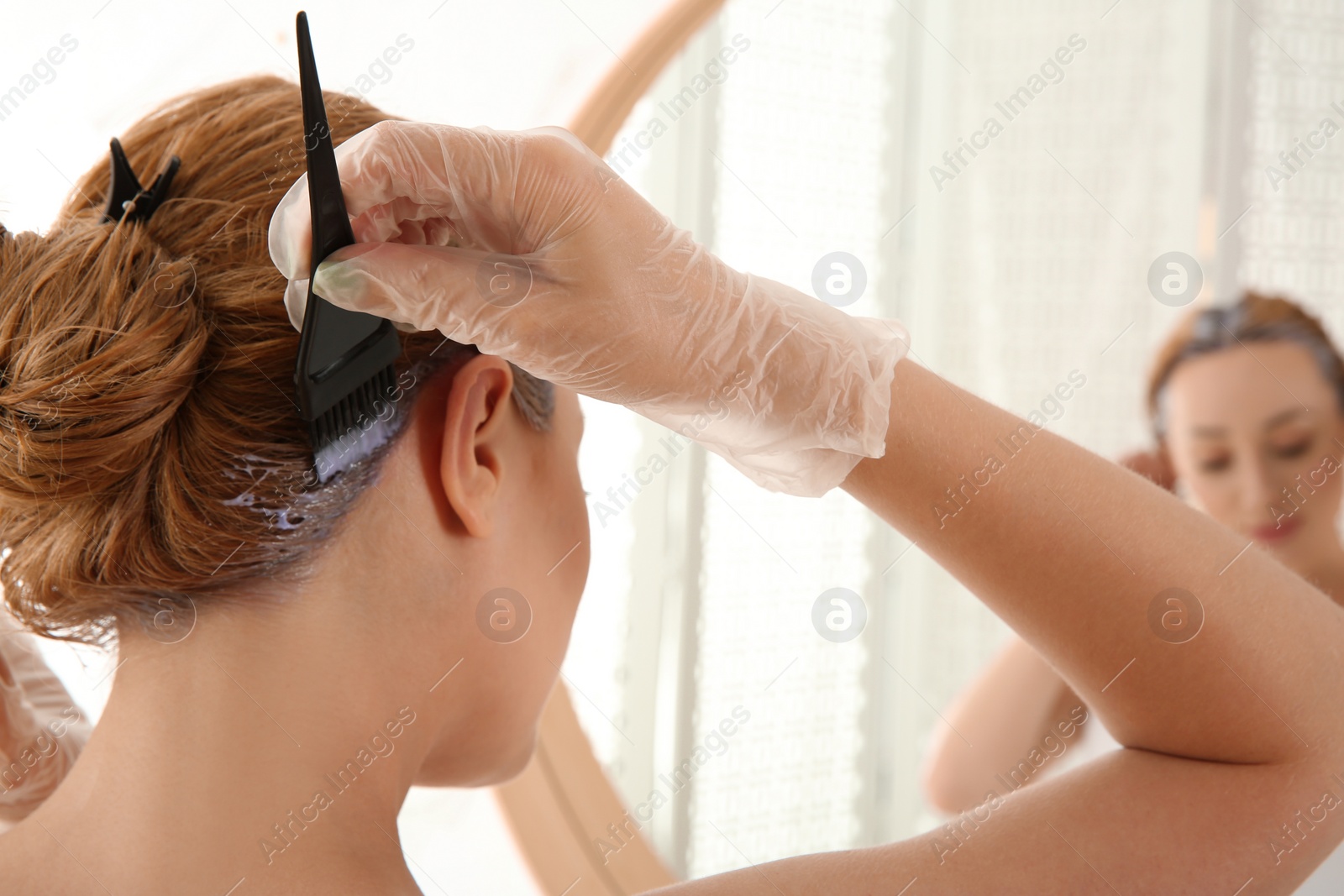 Photo of Young woman applying hair dye on roots in front of mirror at home, closeup