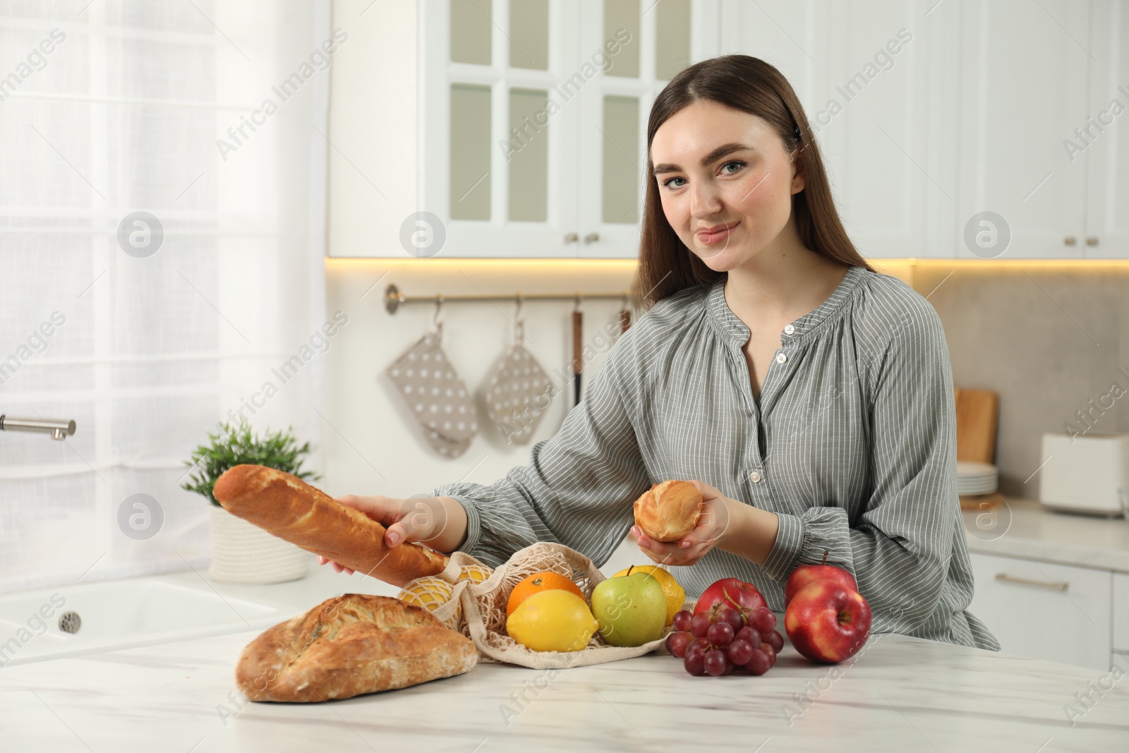 Photo of Woman with baguettes and string bag of fresh fruits at light marble table in kitchen