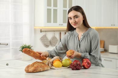 Woman with baguettes and string bag of fresh fruits at light marble table in kitchen