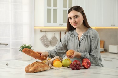 Photo of Woman with baguettes and string bag of fresh fruits at light marble table in kitchen