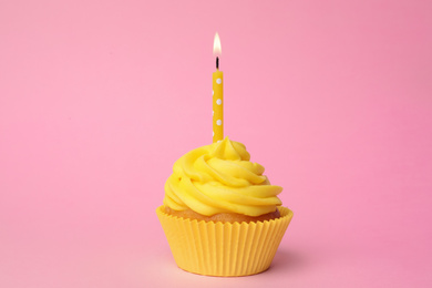 Delicious birthday cupcake with yellow cream and burning candle on pink background