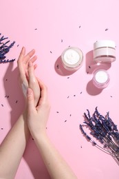 Woman applying hand cream and lavender flowers on pink background, top view