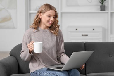 Beautiful woman with blonde hair holding cup and laptop on sofa indoors. Space for text