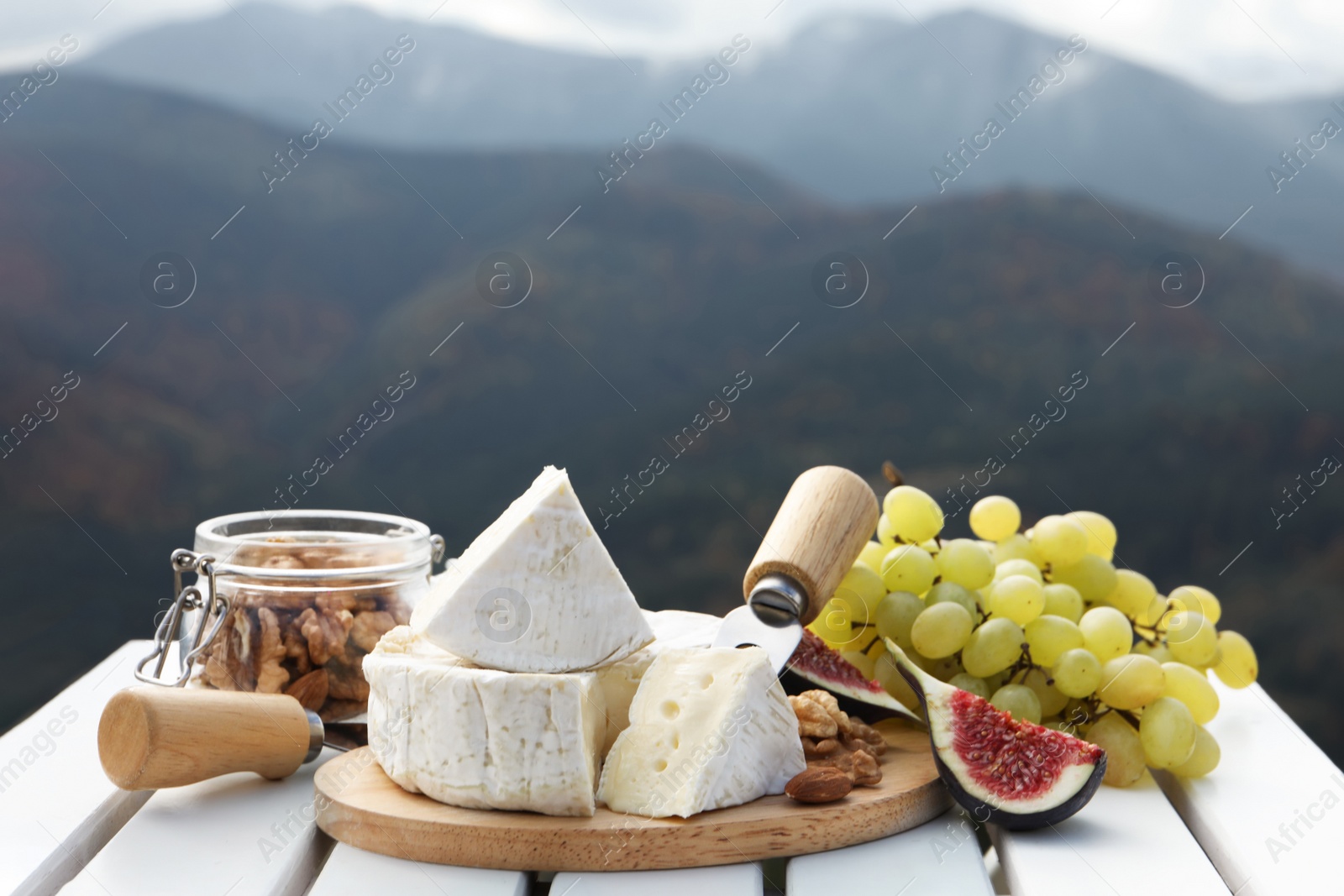 Photo of Delicious cheese, nuts and fruits on white wooden table against mountain landscape