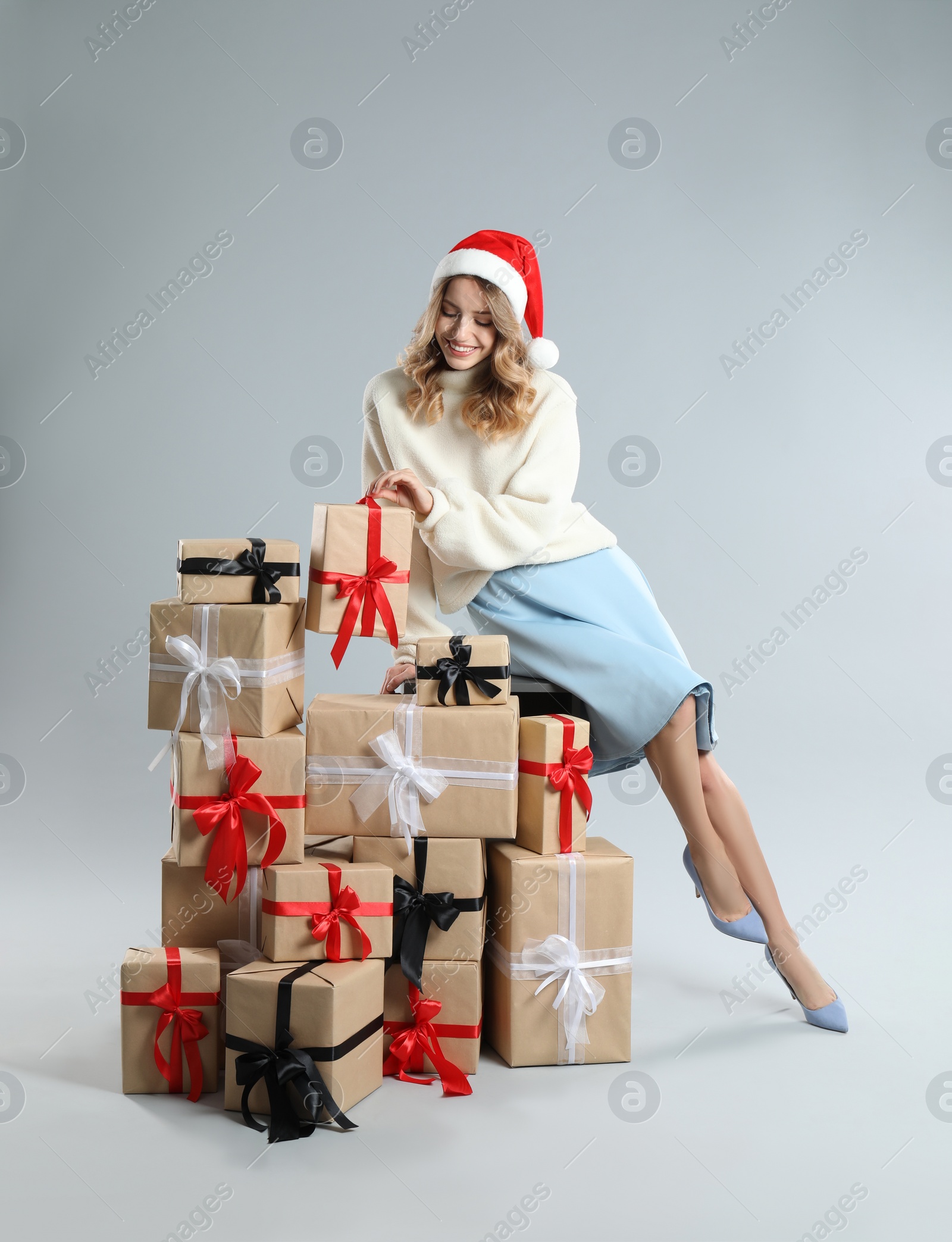Photo of Beautiful young woman in Santa hat with pile of Christmas presents on light grey background