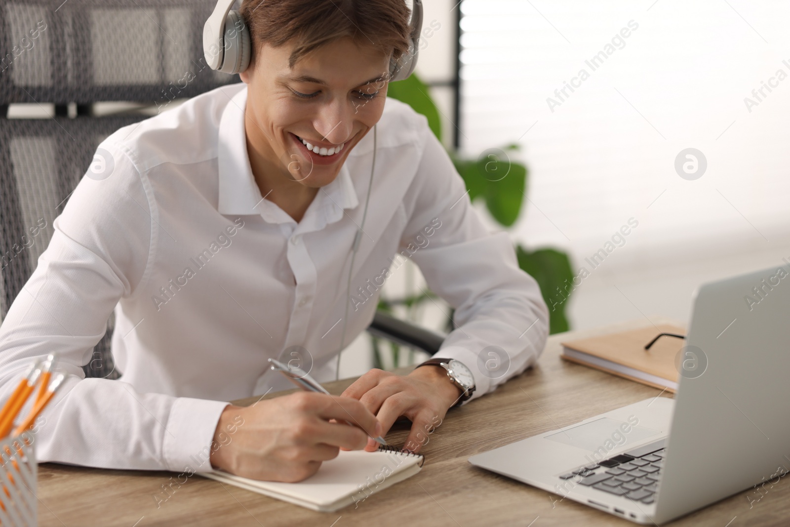 Photo of Man in headphones taking notes during webinar at wooden table indoors