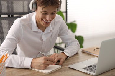 Man in headphones taking notes during webinar at wooden table indoors