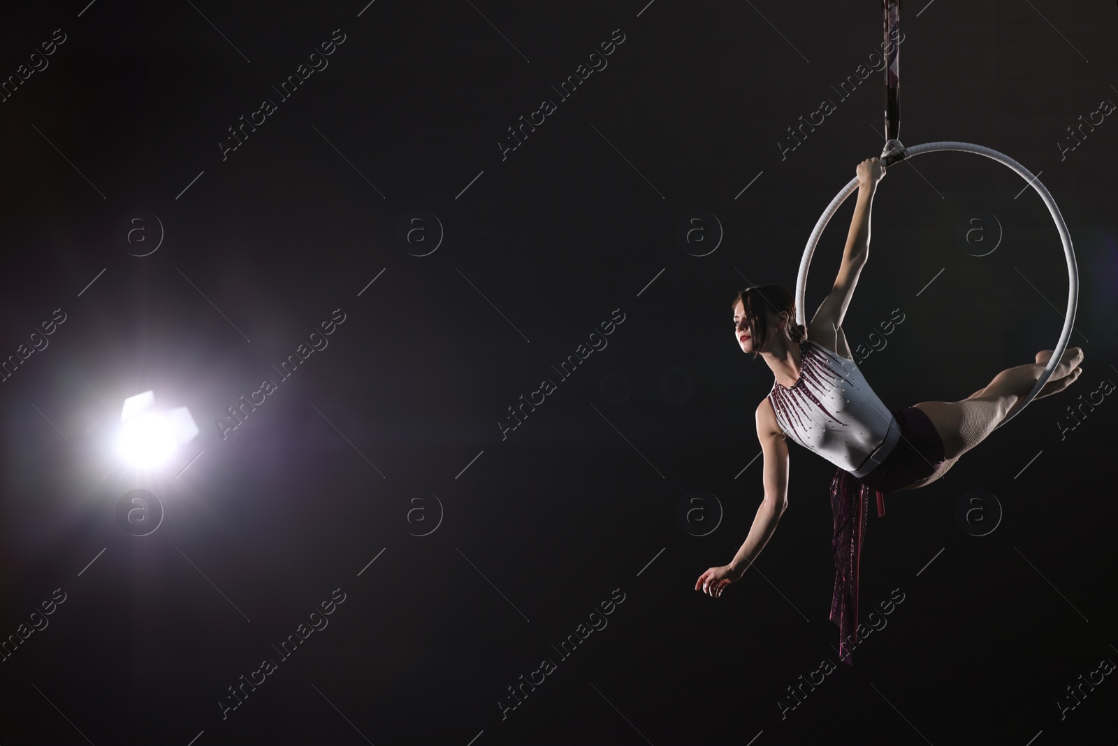 Photo of Young woman performing acrobatic element on aerial ring against dark background. Space for text