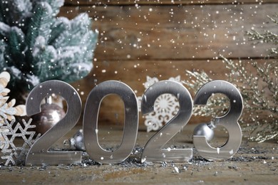 Photo of Snow falling onto silver number 2023, confetti and festive decor on wooden table. Happy New Year