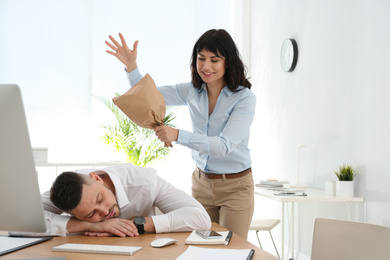 Photo of Young woman popping paper bag behind her sleeping colleague in office. April fool's day