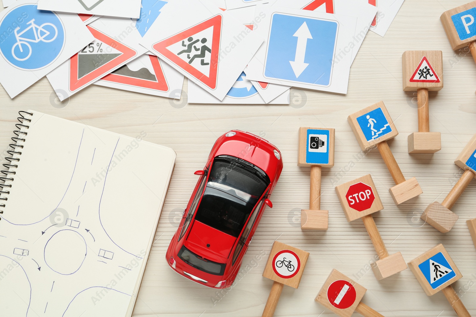 Photo of Many different road signs, notebook with sketch of roundabout and toy car on white wooden background, flat lay. Driving school