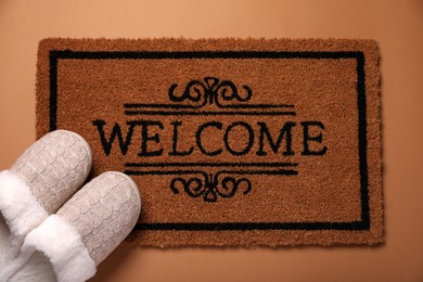 Doormat with word Welcome and slippers on brown background, flat lay