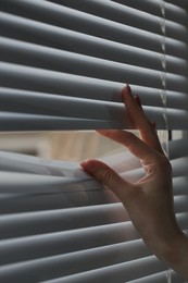 Photo of Woman separating slats of white blinds indoors, closeup